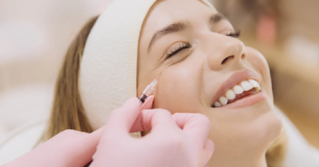 A smiling woman with a headband receives a Botox injection near her eye from a professional wearing pink gloves in St. Charles, IL